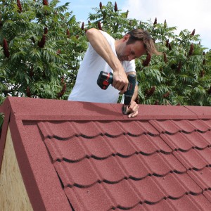 Mann montiert rote Easypan Metalldachpfanne in Ziegeloptik mit Akkuschrauber.