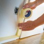 Hände kleben beiges LUX-TOOLS Malerkrepp 30 mm an Wand für präzise Farbkanten bei Renovierungsarbeiten.