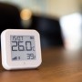 Weißer Shelly Temperatur- und Feuchtigkeitssensor Plus H&T G3 auf einem Tisch, der 26 Grad und 39% Luftfeuchtigkeit anzeigt.