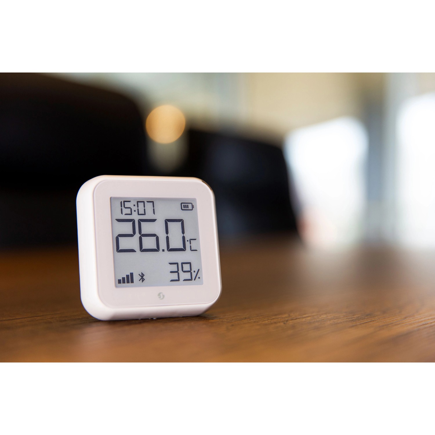 Weißer Shelly Temperatur- und Feuchtigkeitssensor Plus H&T G3 auf einem Tisch, der 26 Grad und 39% Luftfeuchtigkeit anzeigt.