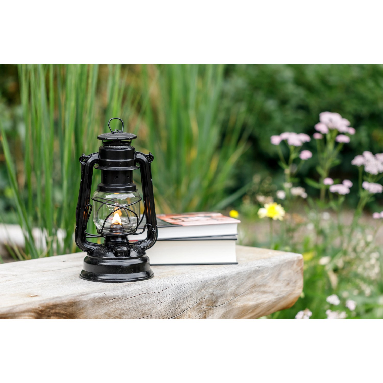 Mattschwarze Feuerhand Sturmlaterne Baby Special 276 auf Holzbank mit Büchern und Blumen im Hintergrund.