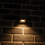 Graue, eckige LED Außenwandleuchte mit warmweißem Licht an einer Backsteinwand.