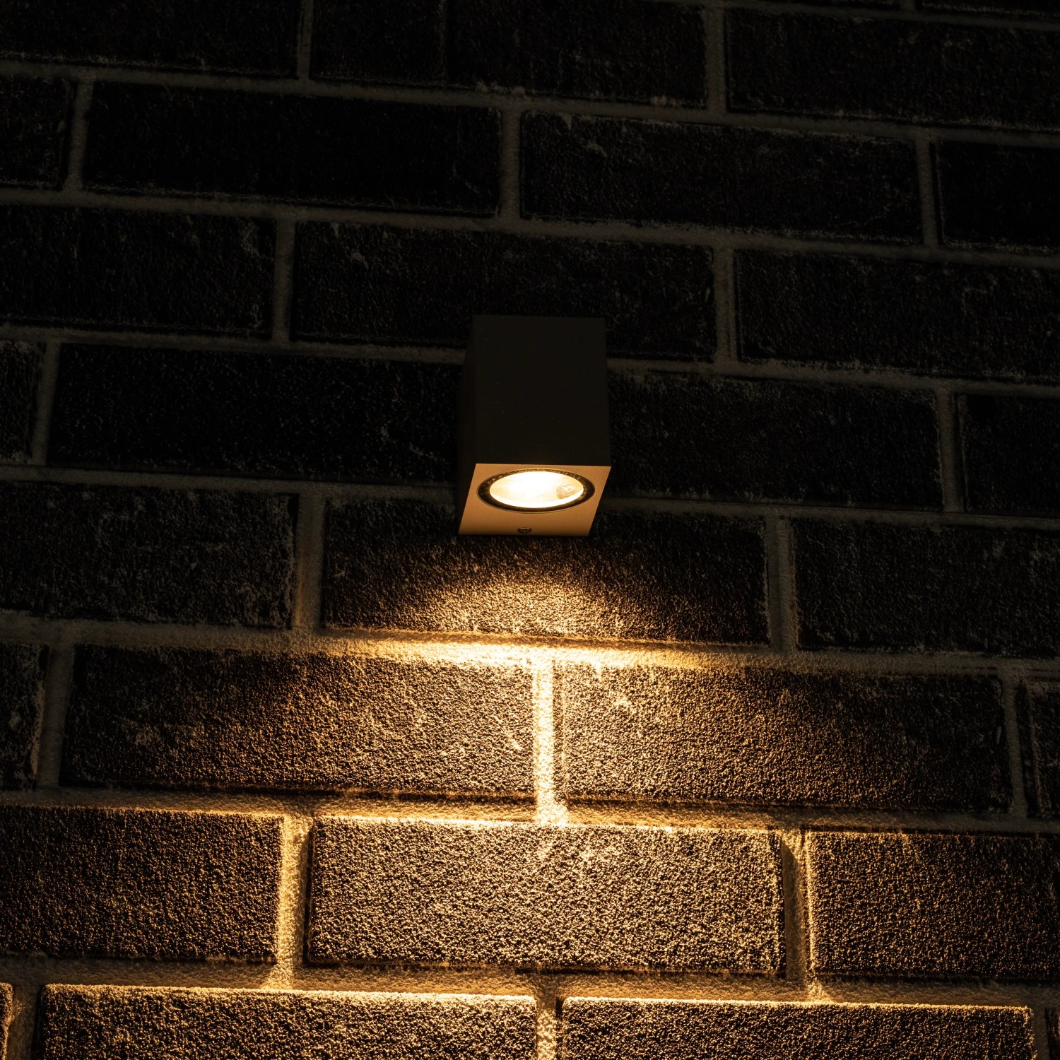 Graue, eckige LED Außenwandleuchte mit warmweißem Licht an einer Backsteinwand.