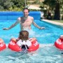 Drei Kinder mit roten Freds Swim Academy Schwimmreifen im Pool mit Erwachsenem.