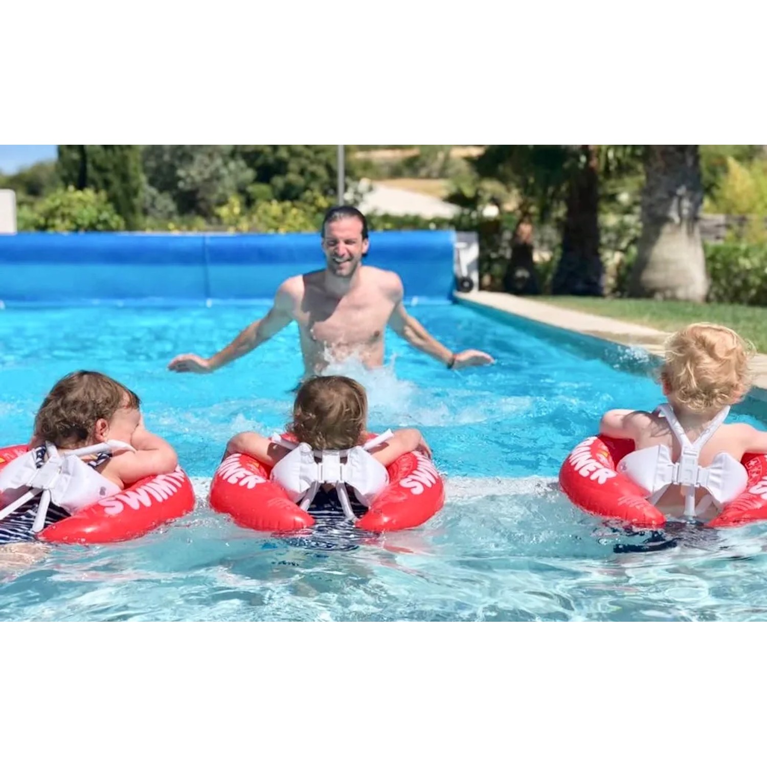 Drei Kinder mit roten Freds Swim Academy Schwimmreifen im Pool mit Erwachsenem.