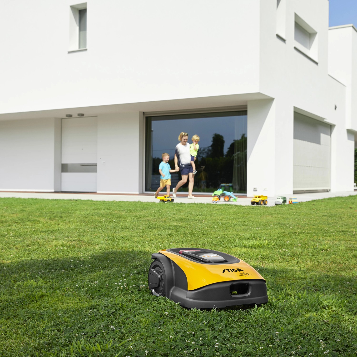 Gelb-schwarzer Stiga Mähroboter G 600 auf Rasen vor modernem Haus mit Familie.