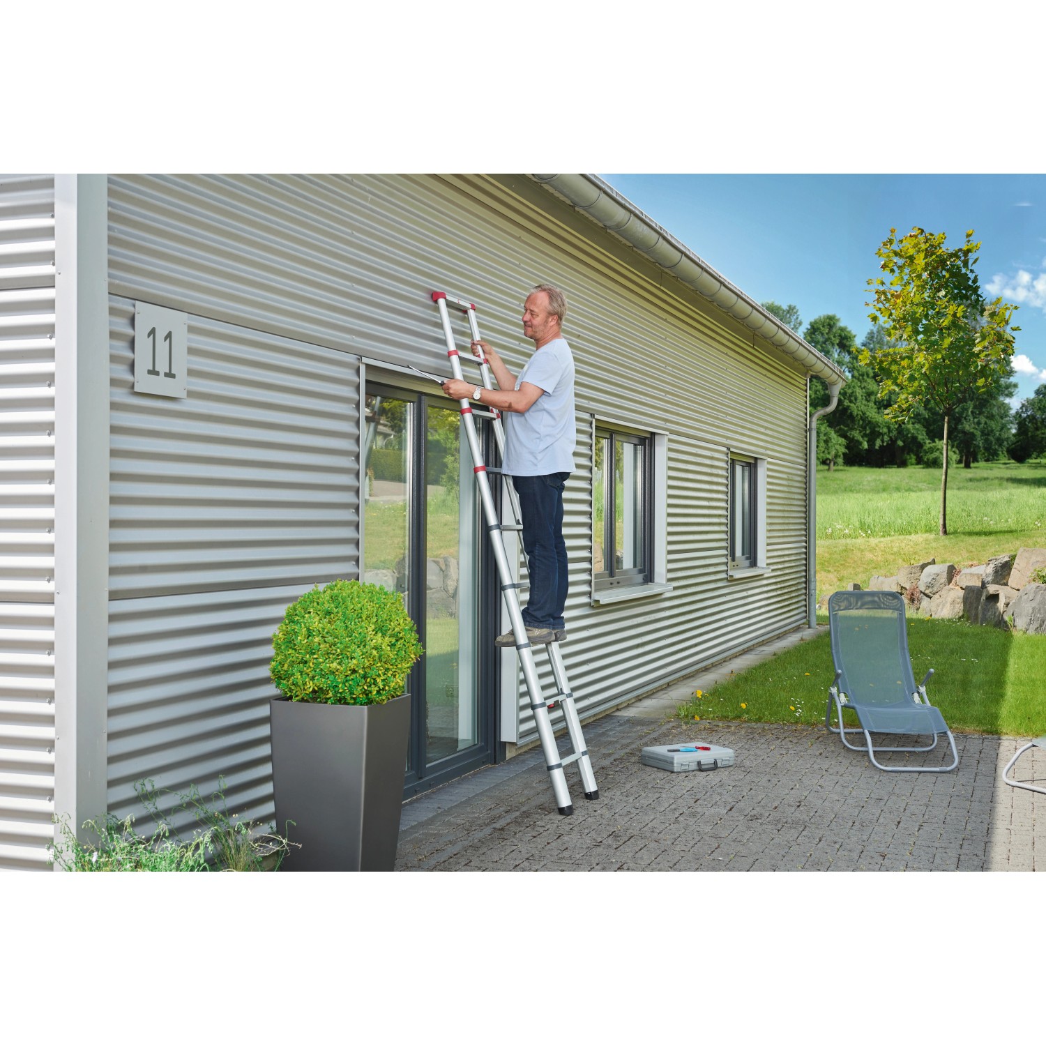 Mann auf Hailo Alu-Teleskopleiter FlexLine 260 (9-stufig) vor Hauswand. Leiter bis 150 kg belastbar.