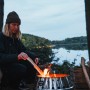 Petromax Atago Grill-Set mit Frau am Lagerfeuer im Freien. Feuerschale mit Grillspießen.