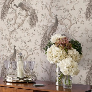 Laura Ashley Vliestapete Belvedere Soft Truffle mit Pfauenmuster und Blumen auf beiger Wand.