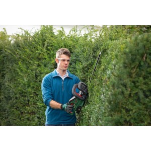 Mann schneidet Hecke mit Bosch Akku-Heckenschere UniversalHedgeCut 18-55. Grünes Gerät, Schutzbrille, Handschuhe.