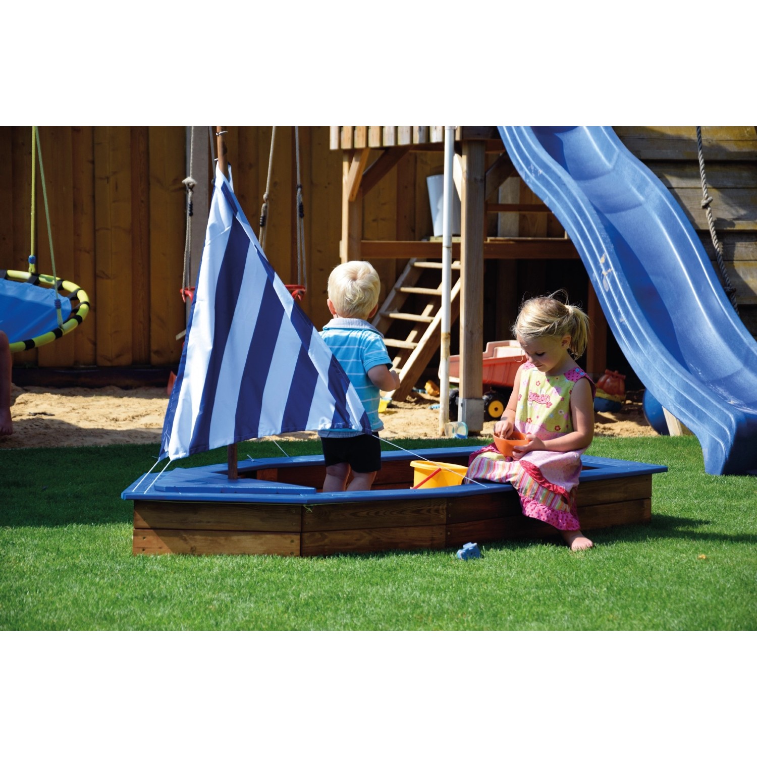 Sandkasten im Schiff-Design mit blau-weißem Segel und spielenden Kindern im Garten.