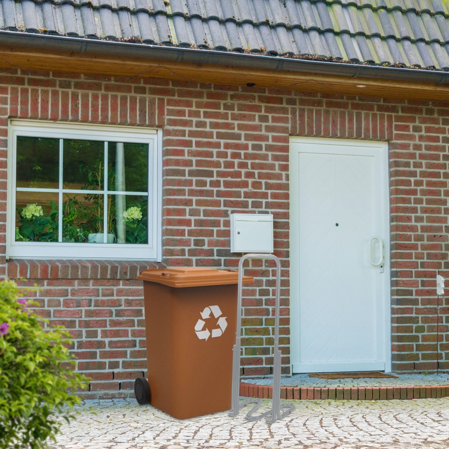 Graue Relaxdays Müllpresse aus Stahl, manuell, neben brauner Mülltonne vor Haus. Reduziert Abfallvolumen effektiv.