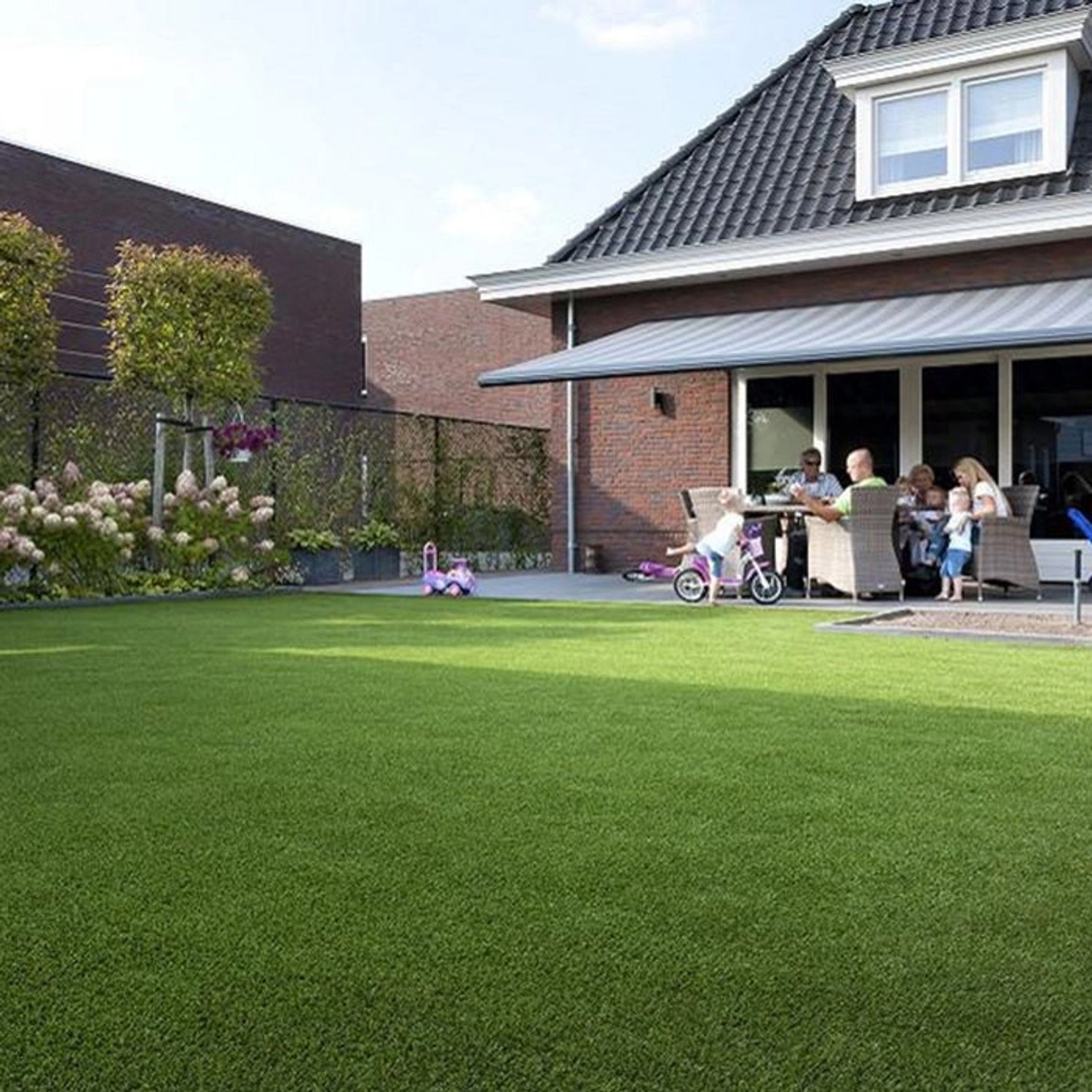 Sonniger Garten mit Karat Kunstrasen Nantes, 40mm hoch, UV-beständig. Familie sitzt auf der Terrasse.