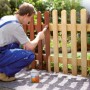 Mann streicht Zaun im Garten; graues LUX-TOOLS Abdeckvlies schützt den Boden vor Farbspritzern. Rutschfest & wasserundurchlässig.
