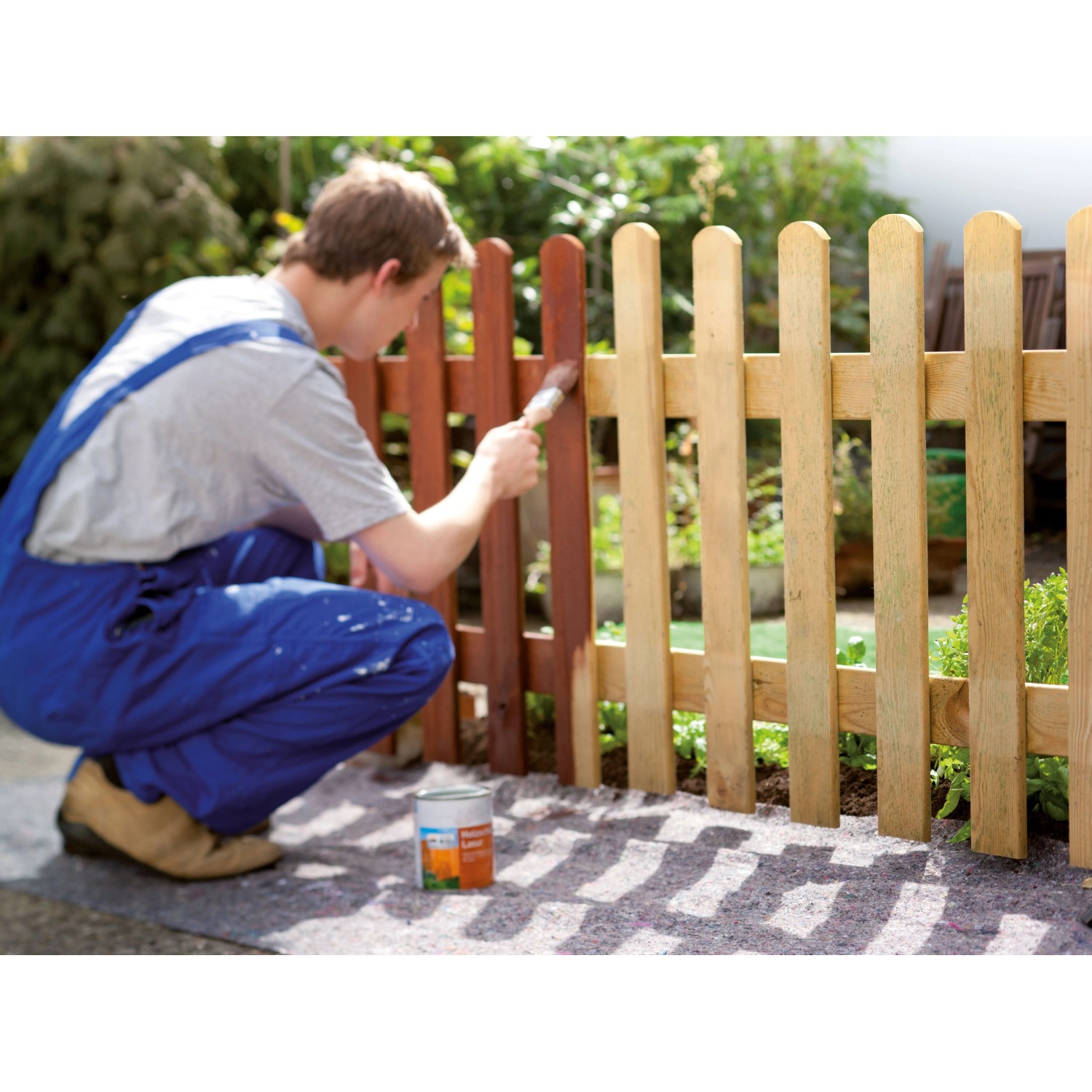 Mann streicht Zaun im Garten; graues LUX-TOOLS Abdeckvlies schützt den Boden vor Farbspritzern. Rutschfest & wasserundurchlässig.