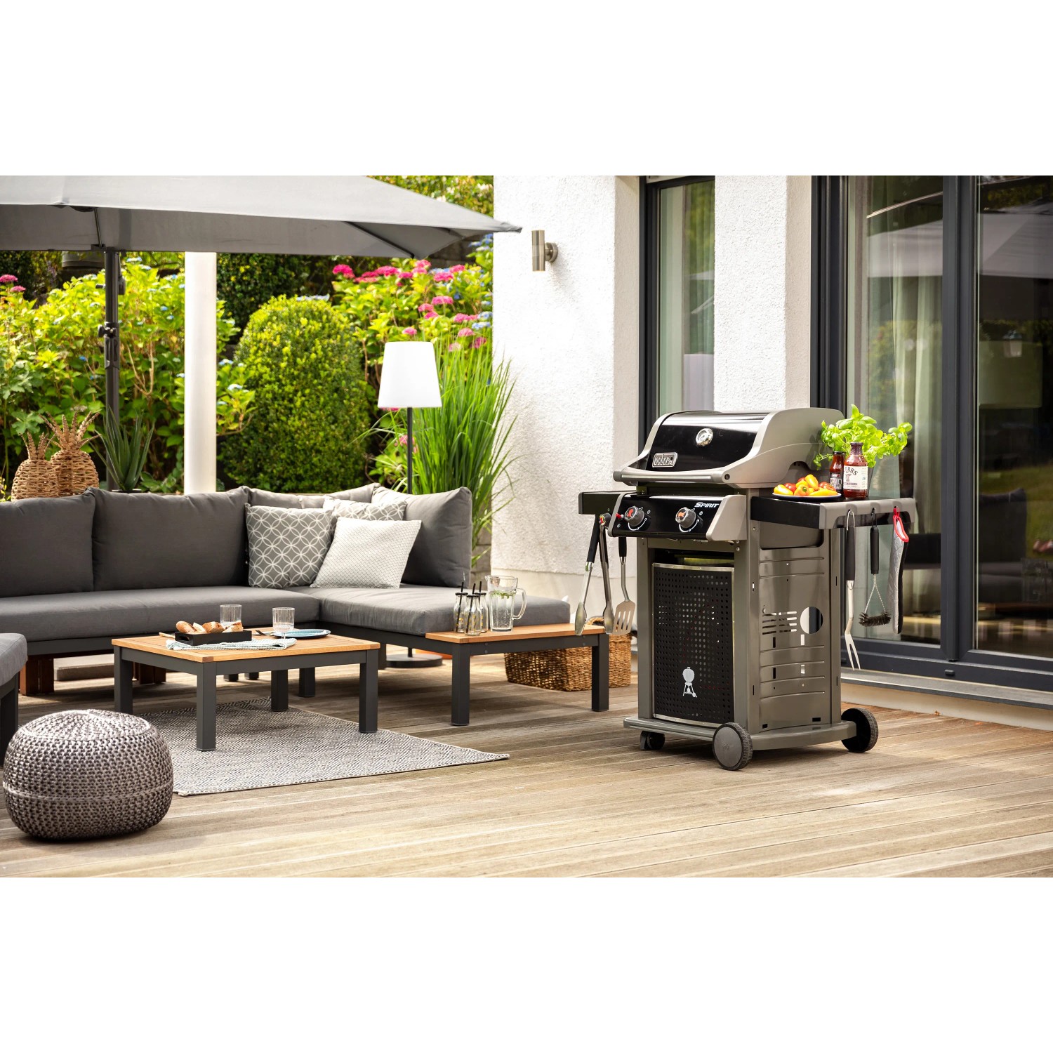 Gartenlounge mit grauer Eckcouch, Tisch und Grill auf einer Holzterrasse.