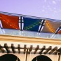 Mehrere gespannte, viereckige Sonnensegel in Rot, Blau, Grau mit Lichterketten über einer Terrasse. AMANKA Schwerlast Befestigung.