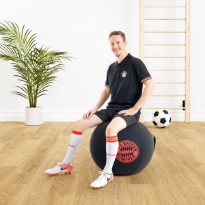 Mann sitzt auf schwarzem Sitting Ball Hocker FCB (120 l) mit Bayern München Logo.