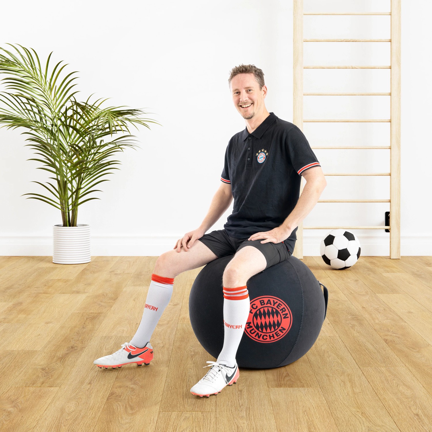 Mann sitzt auf schwarzem Sitting Ball Hocker FCB (120 l) mit Bayern München Logo.