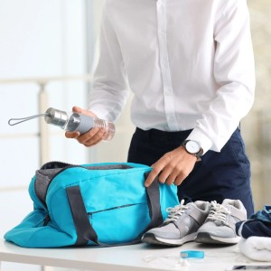 Person packt blaue Sporttasche mit Wasserflasche und Turnschuhen; für ordentliche Aufbewahrung unterwegs.