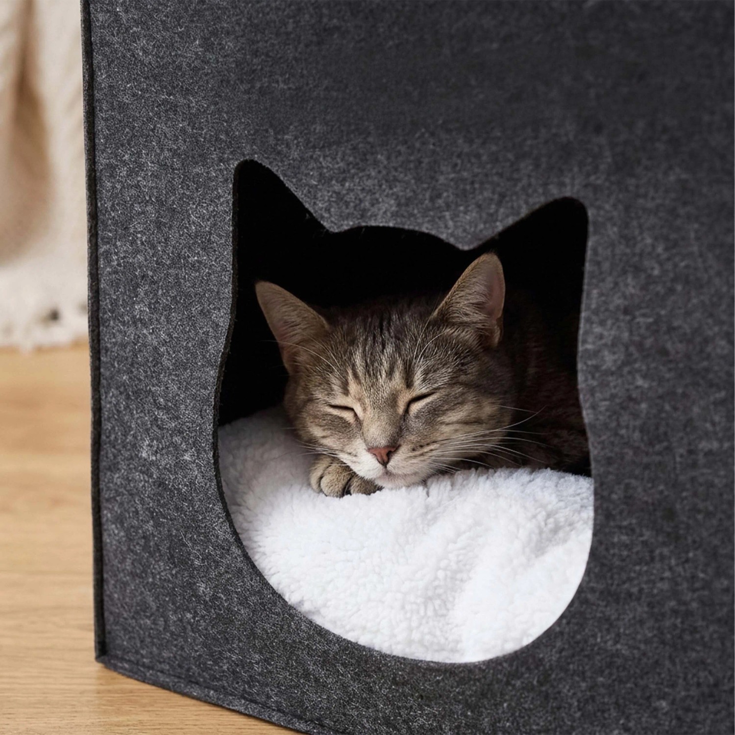 Dunkelgraue Filz Katzenhöhle mit Katzenkopf-Eingang und flauschigem Kissen, in der eine Katze schläft.