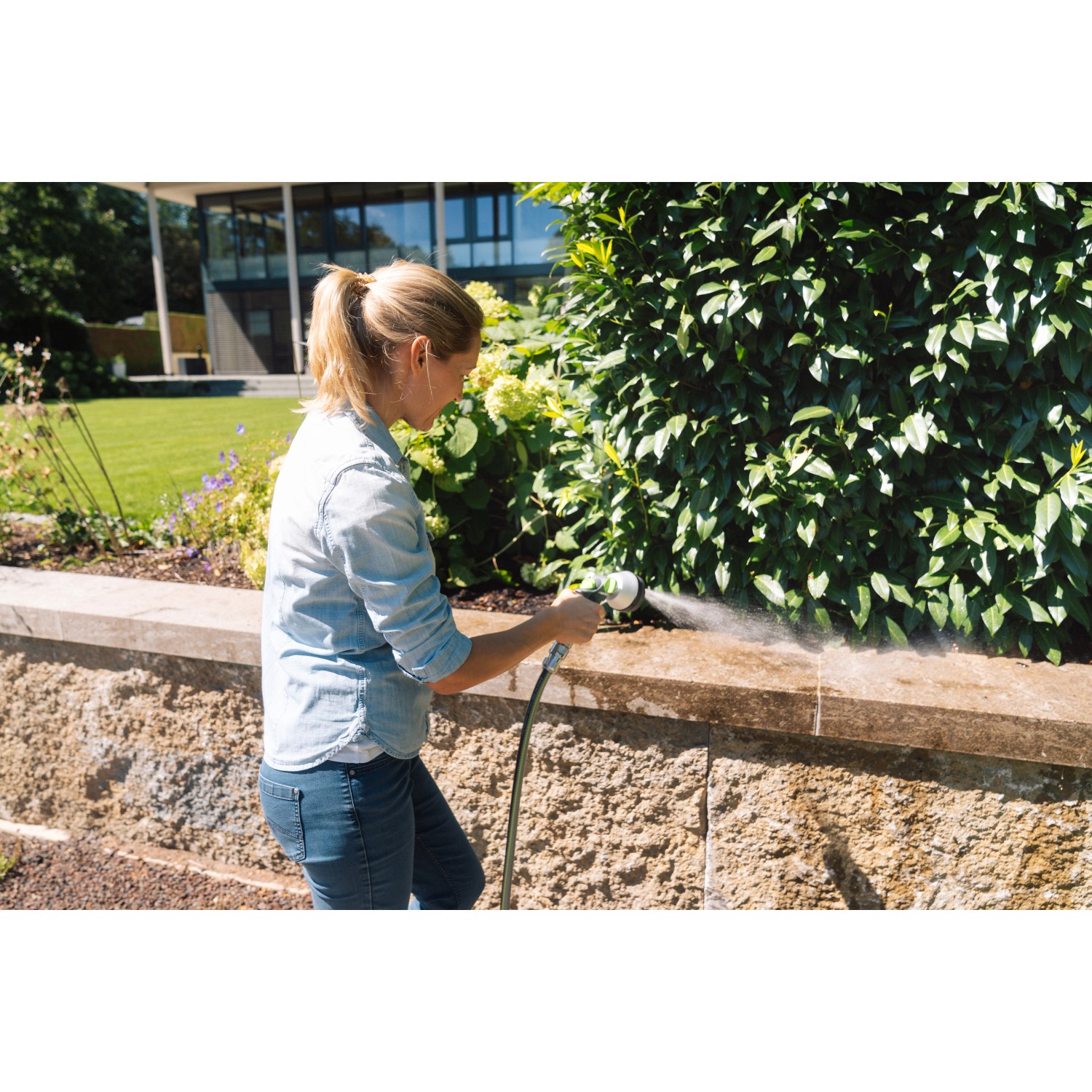 Frau nutzt graue Geka Multibrause Plus 3S Gartenspritze zur Reinigung einer Mauer im Garten.