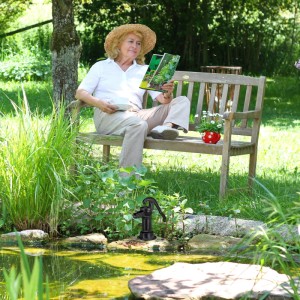 Dekorative, schwarze Gartenpumpe aus Gusseisen am Teich, Frau liest auf Bank im Hintergrund.