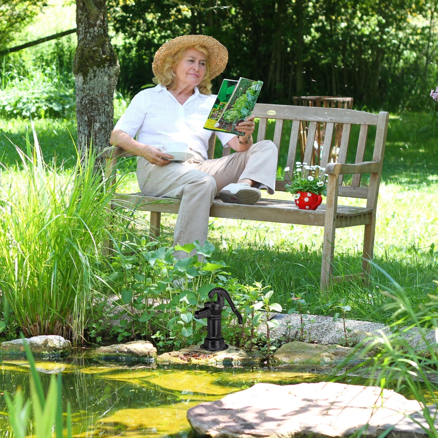 Dekorative, schwarze Gartenpumpe aus Gusseisen am Teich, Frau liest auf Bank im Hintergrund.