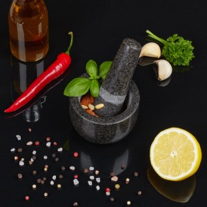 Grauer Granit Mörser mit Stößel, umgeben von frischen Zutaten wie Basilikum, Chili, Knoblauch und Zitrone. Ideal für Pesto.