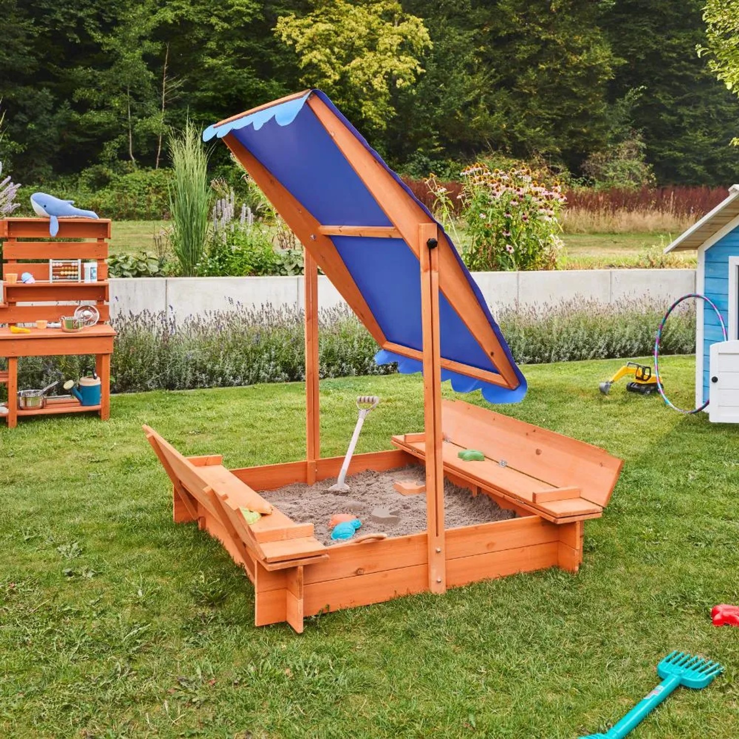 Home Deluxe Sandkasten Buddelkiste mit blauem Dach und Sitzbänken im Garten.