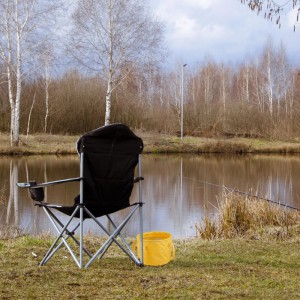 Gelber Falteimer aus Kunststoff, zusammenklappbar, am Seeufer neben Campingstuhl. Ideal für Camping & Angeln.