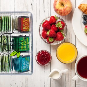 Transparente Relaxdays Teebox mit 6 Fächern für Teebeutel auf Holztisch, neben Frühstück mit Obst und Saft.