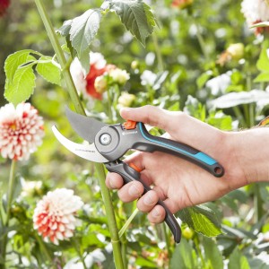 Hand hält Gardena Gartenschere B/M vor blühendem Garten. Bypass-Schere mit blau-grauem Griff.