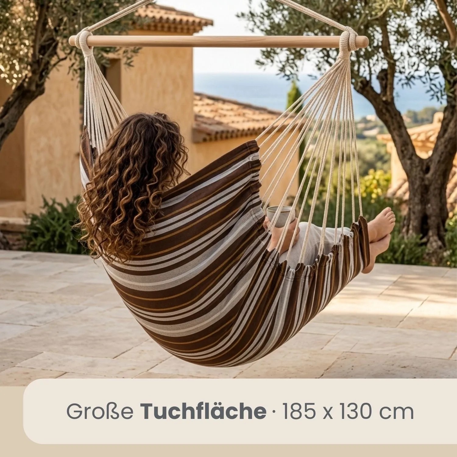 Gestreifter AMANKA Hängesessel aus Baumwolle mit Holzstab, in dem eine Person auf einer Terrasse entspannt. Ideal für Garten & Balkon.