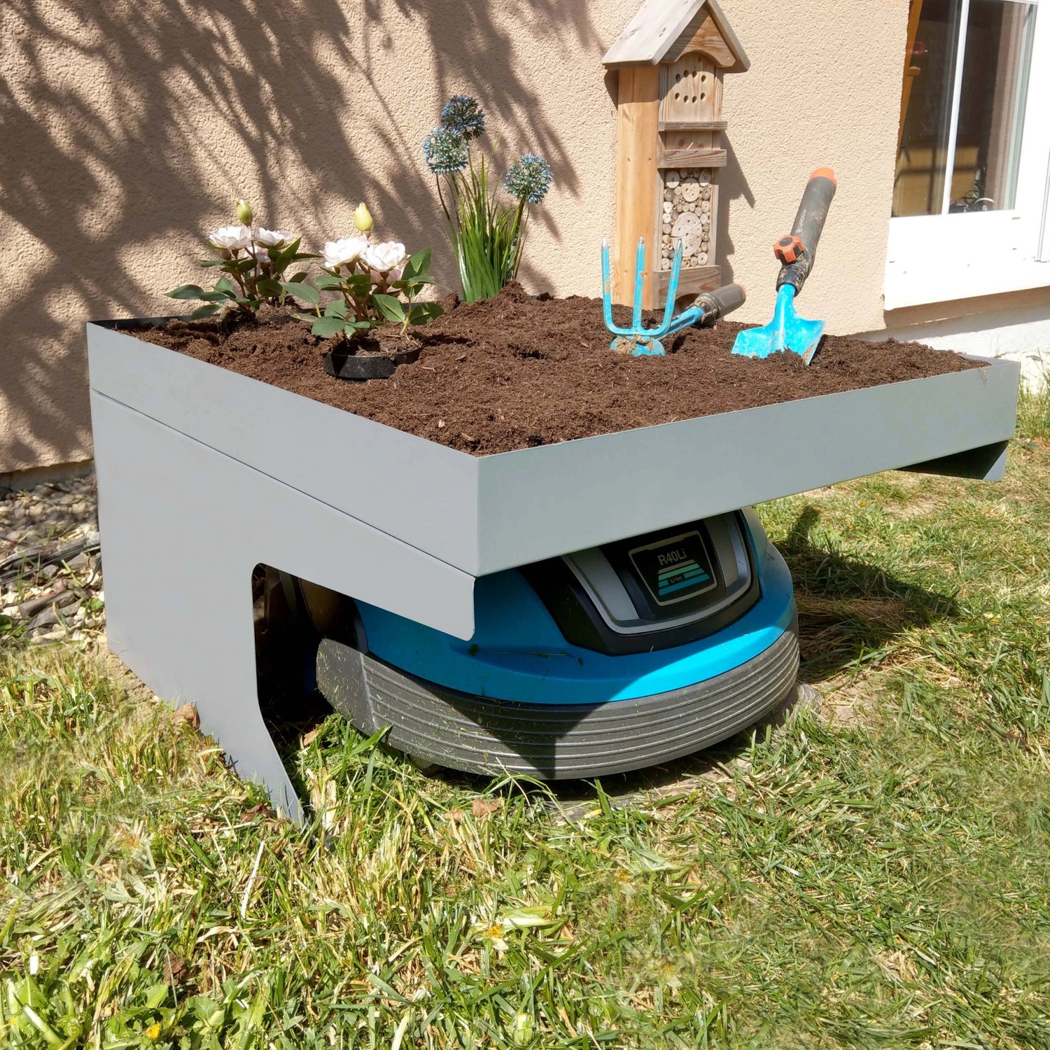 Graue MCW Mähroboter-Garage G60 mit Pflanzkasten und Mähroboter im Garten.