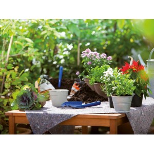 Graues Abdeckvlies auf Holztisch im Garten, schützt bei Umtopfarbeiten mit Pflanzen, Erde und Gartengeräten.