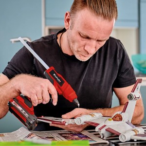 Mann benutzt Einhell Akku-Heißklebepistole TE-CG 18 Li zum Basteln an Modell. Rotes Heißklebegerät mit Akku.