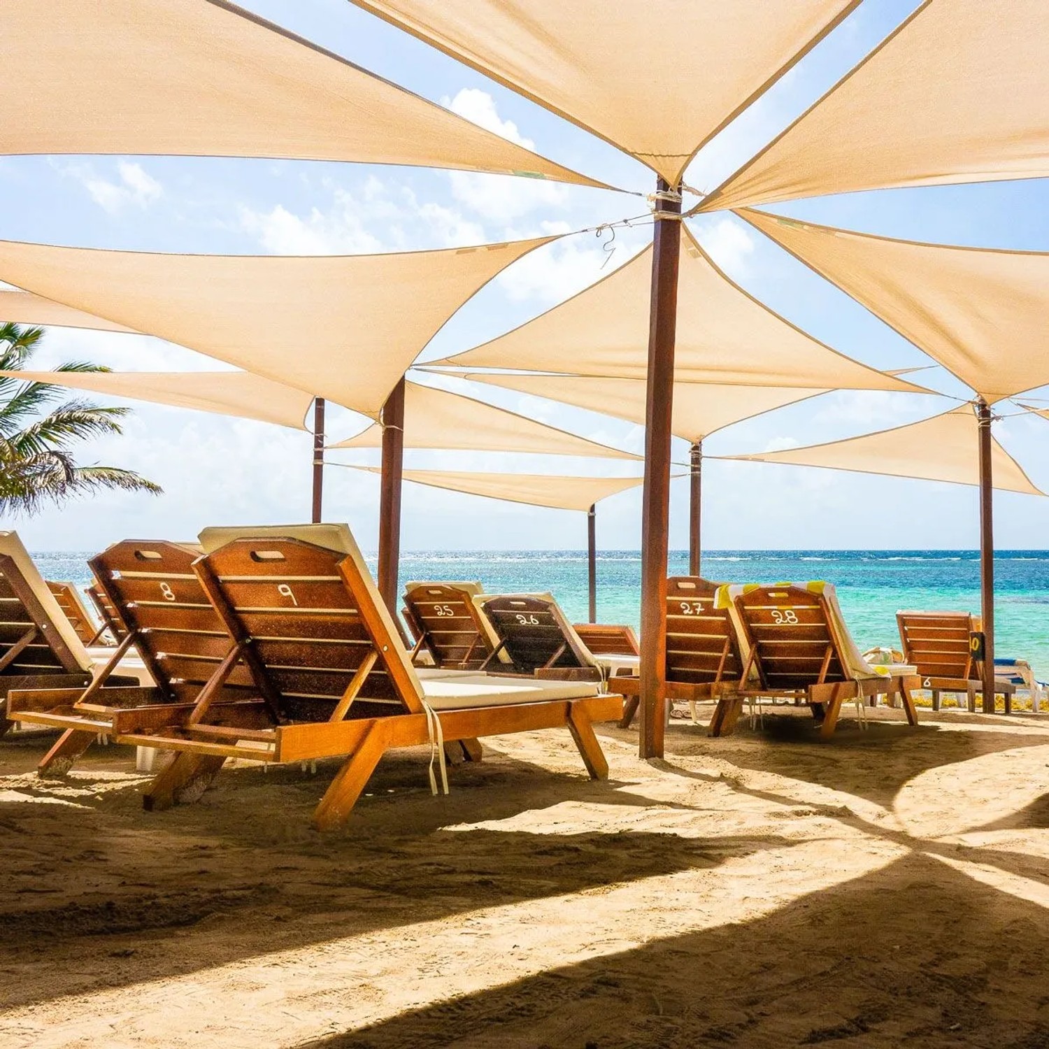 Helle dreieckige Sonnensegel spenden Schatten über Liegestühlen am Strand. Ideal für Schwerlast Sonnensegel Befestigung.