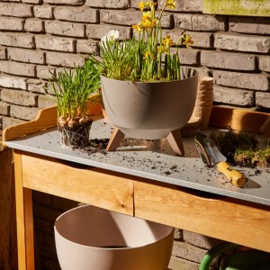 Beiges Siena Garden Pflanzgefäß Oslo (Ø 40 cm) mit Holzfüßen und Bepflanzung auf einem Gartentisch.