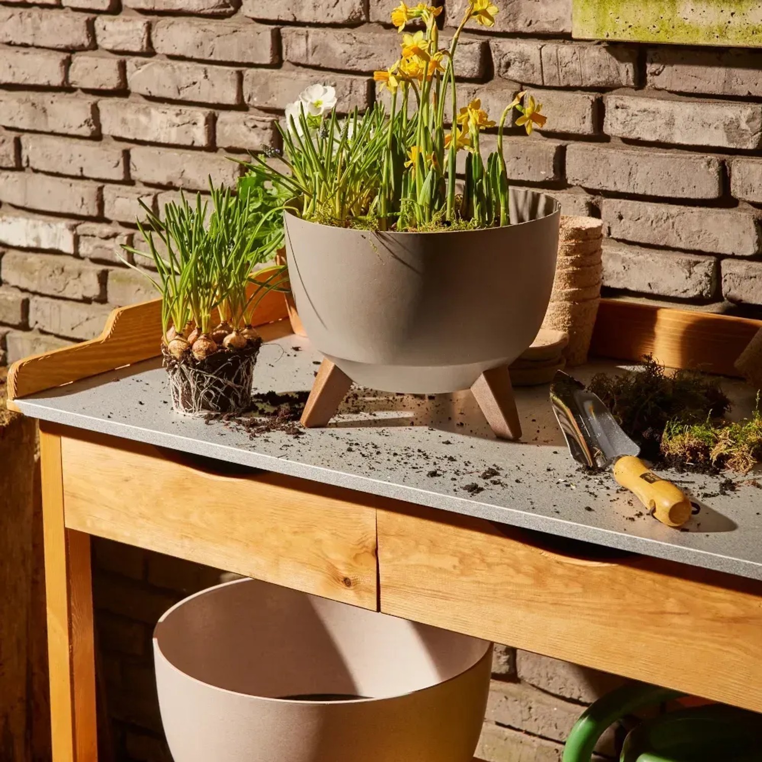 Beiges Siena Garden Pflanzgefäß Oslo (Ø 40 cm) mit Holzfüßen und Bepflanzung auf einem Gartentisch.