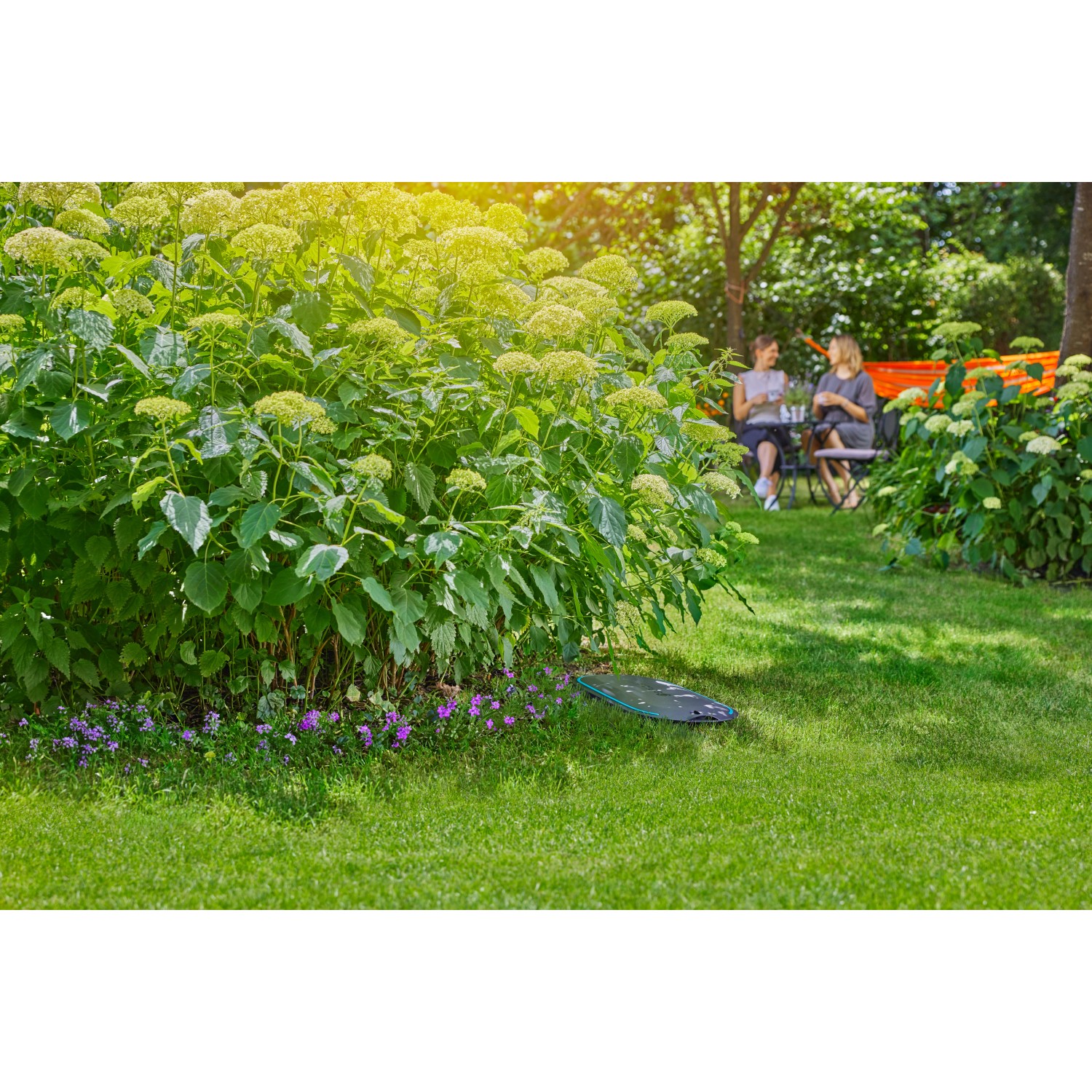 Gardena Undercover Schlauchbox RollUp M im Garten unter einem Busch. Im Hintergrund sitzen zwei Frauen.