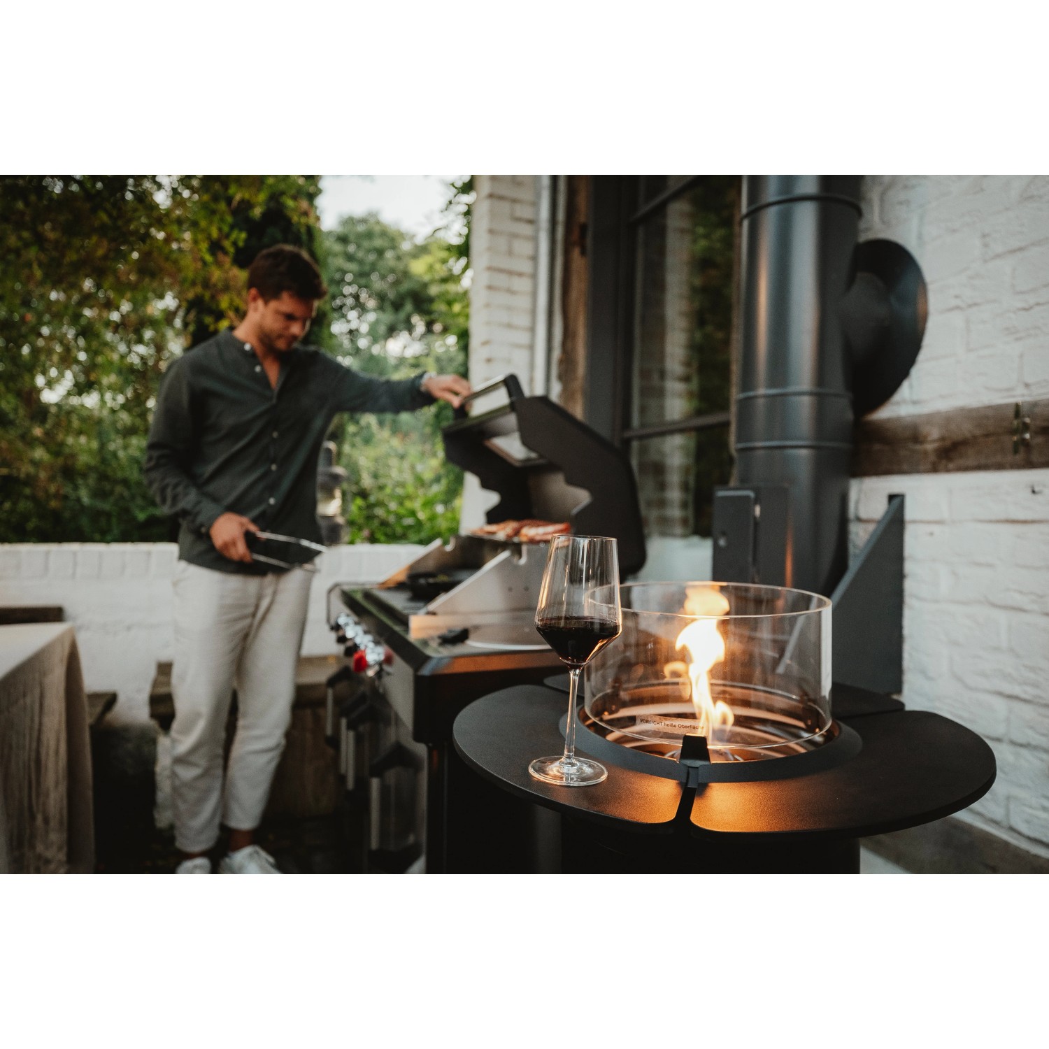 Enders Gas-Terrassenfeuer Nova, schwarz, mit Glaszylinder und Ablage, im Hintergrund ein Grill.