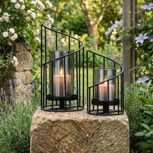 Zwei schwarze Eisen-Windlichter mit Glaszylindern und brennenden Kerzen im Garten. Perfekt als Deko-Set.