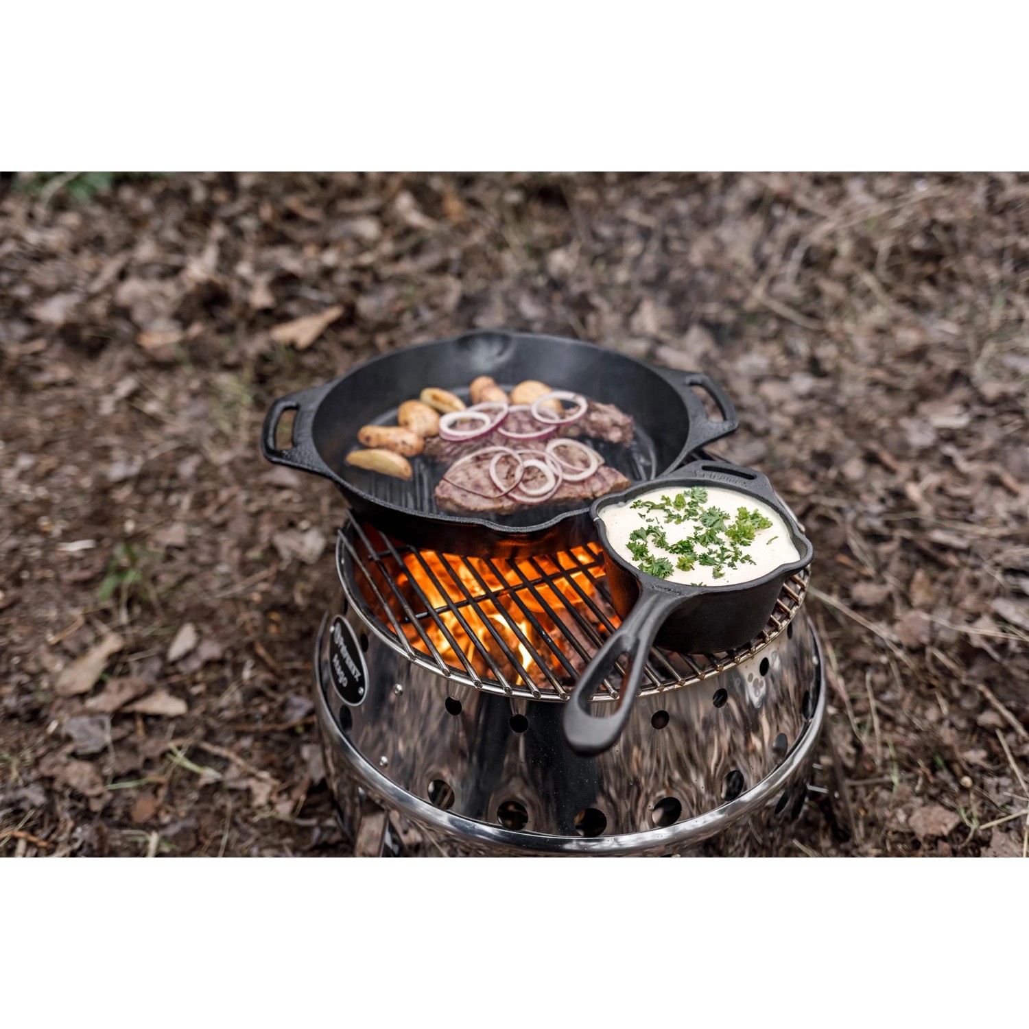Petromax Atago Grill-Set mit Grillspießen beim Outdoor-Kochen mit Pfanne und Topf über dem Feuer.