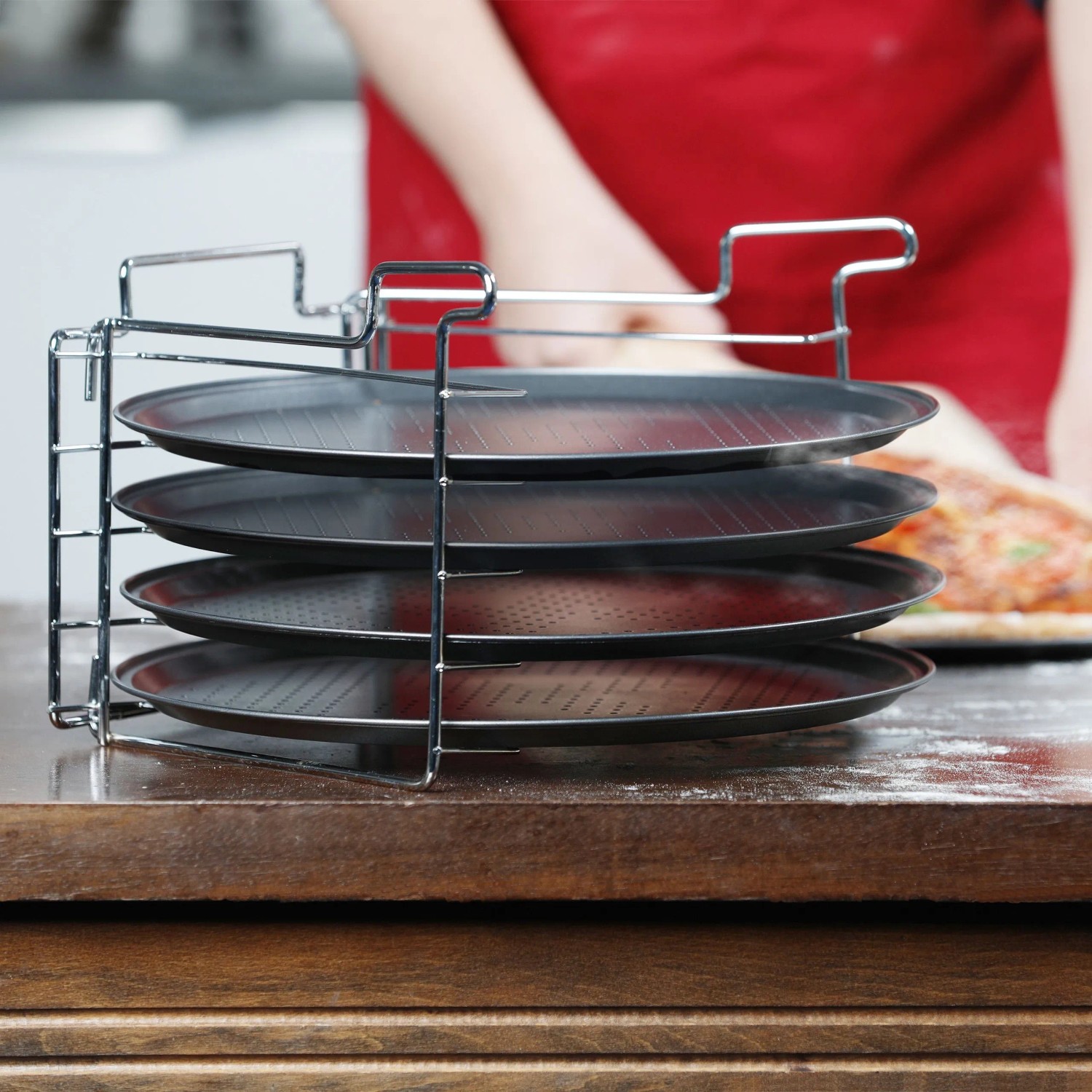 Pizzablech 4er Set mit Ständer: Vier antihaftbeschichtete, gelochte Backbleche aus Eisen im Metallgestell auf Holztisch. Für Pizza.