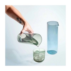 Hand gießt Wasser aus graugrüner Villeroy & Boch Like Glass Karaffe in Glas mit Eis. Daneben blaue Karaffe. Geschirr.