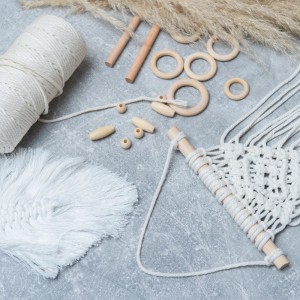 Makramee Set mit weißem Baumwollgarn, Holzperlen, Holzringen und Stäben auf grauem Untergrund für kreatives Basteln.
