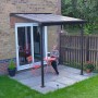 Graues Palram Terrassendach Sierra 230x230 cm mit Frau an Tisch und Stühlen auf Terrasse.