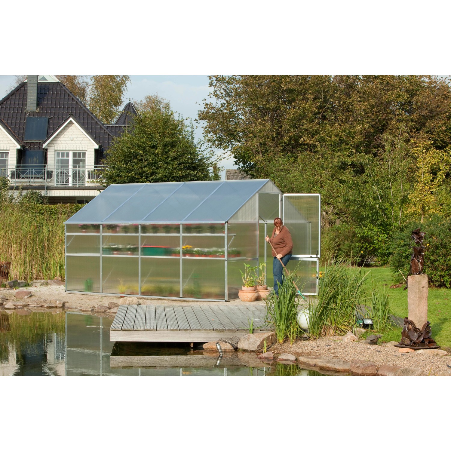 KGT Gewächshaus Alu blank 10 m² mit geöffneter Tür und Bepflanzung im Garten am Teich.