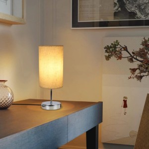 Moderne dimmbare Tischlampe mit hellem Stoffschirm und glänzendem Metallfuß auf einem Schreibtisch. Mit USB-Ladefunktion.